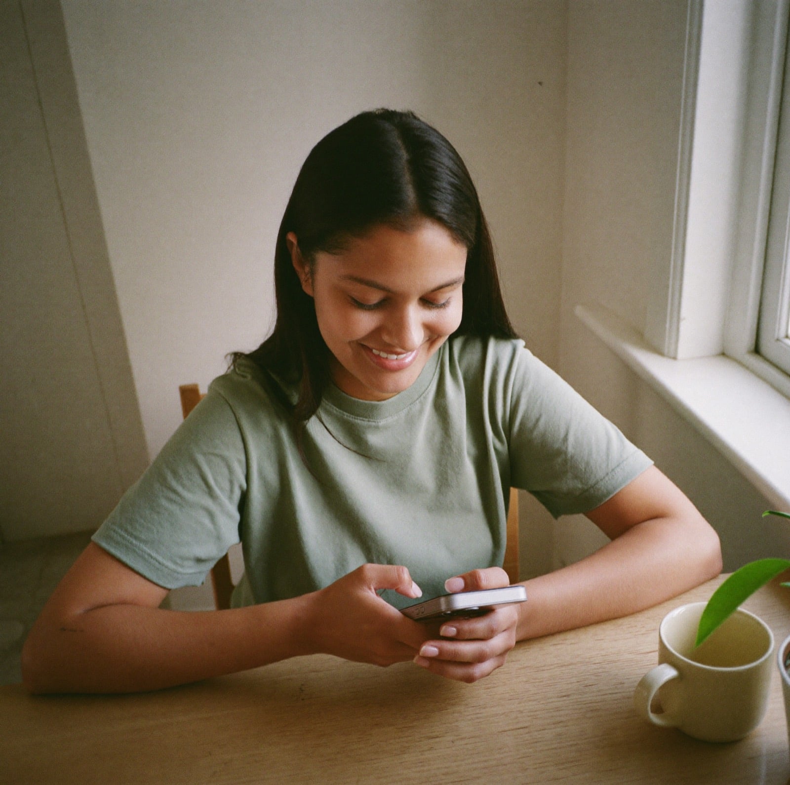 Una joven sonríe mientras contesta en su celular, en la mesa de su cocina, junto a una ventana.